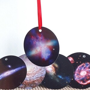 Puede incluir: Cinco adornos redondos con imágenes de diferentes galaxias y nebulosas. Los adornos son negros con imágenes coloridas del espacio. Los adornos cuelgan de una cinta roja.
