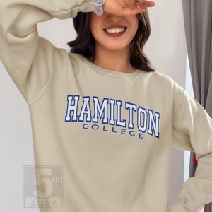 Peut inclure: Un sweat-shirt beige clair avec le texte "HAMILTON COLLEGE" en lettres blanches soulignées de bleu.