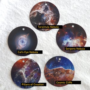 Puede incluir: Cinco adornos redondos con imágenes de diferentes nebulosas. Los adornos están etiquetados como "Nebulosa de la Tarántula", "Nebulosa del Ojo de Gato", "Nebulosa de la Serpiente", "Pilares de la Creación" y "Acantilados Cósmicos".