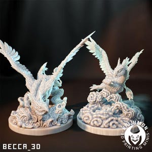 May include: Two white 3D printed miniature figures of mythical creatures with feathered wings, perched on cloud-like bases. The figures are detailed and intricate, with a textured, feathery appearance.