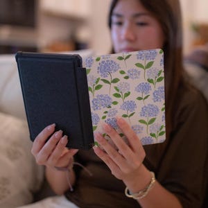 May include: A rectangular e-reader device featuring a protective case with a white background and a repeating pattern of blue hydrangea flowers with green stems and leaves. The back of the case is solid black with a textured, matte finish.
