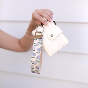 May include: A cream-colored wallet with a snap closure and a matching wristlet strap featuring a pattern of blue hydrangea flowers and green stems, attached by a bronze-toned metal ring and hardware clasp.