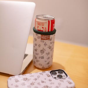 May include: A white floral print can cooler with a green trim holds a red and white can of soda. A matching floral print phone case is next to the cooler. A laptop is in the background. The can cooler has the text "Frederick Family Co."
