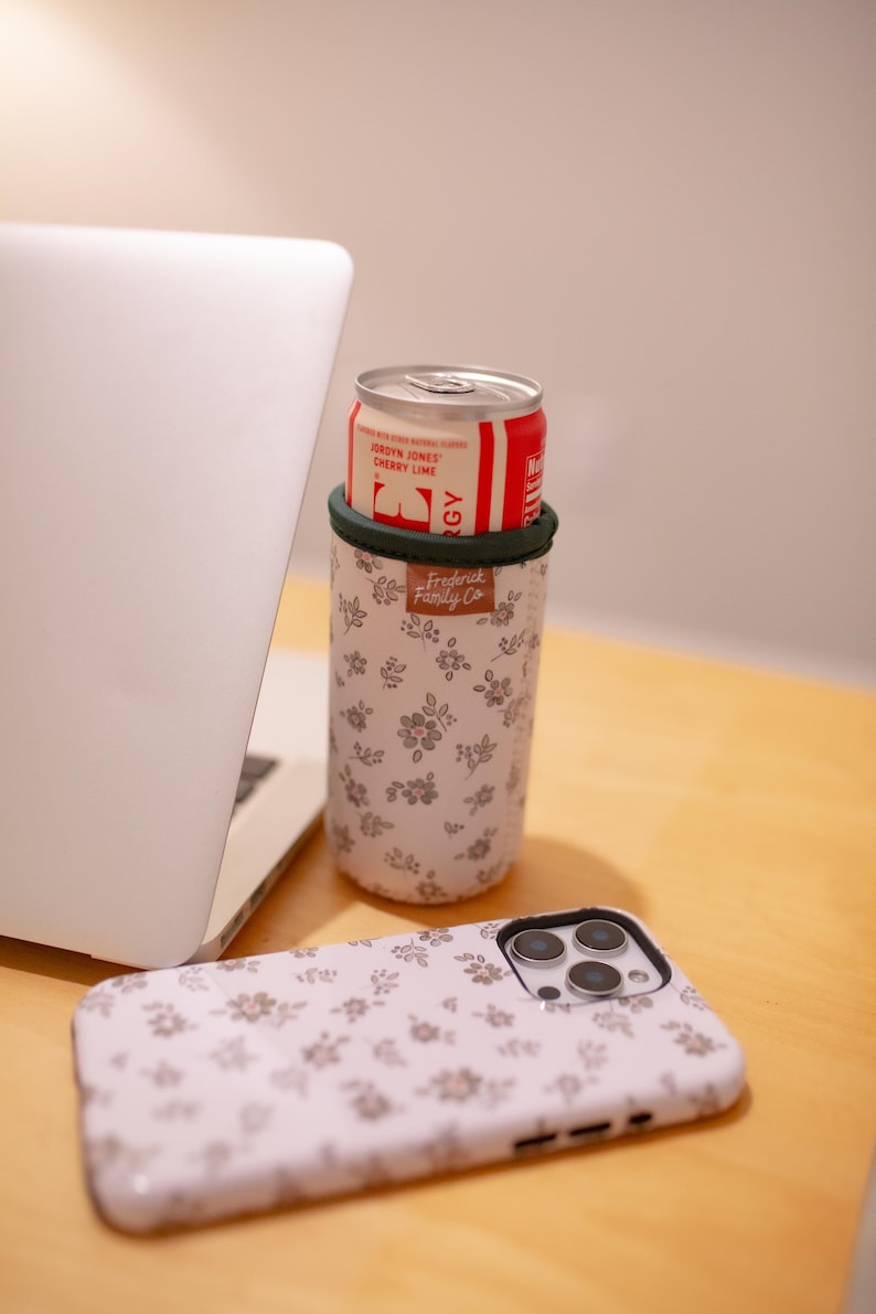 May include: A white floral print can cooler with a green trim holds a red and white can of soda. A matching floral print phone case is next to the cooler. A laptop is in the background. The can cooler has the text "Frederick Family Co."