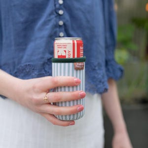 May include: A striped beverage insulator in shades of blue and white, holding a red and white can of Jordyn Jones Cherry Lime. The insulator has a green rim and a brown leatherette tag that reads "Frederic Family Co."