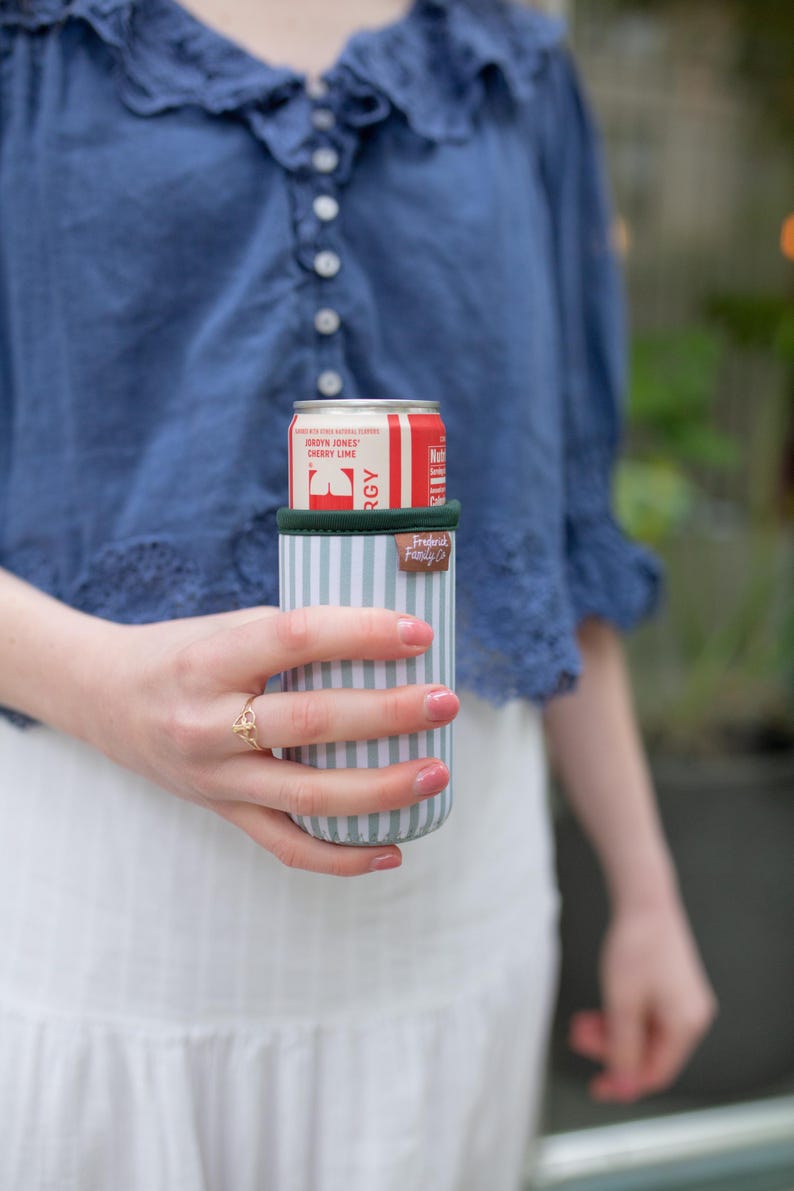 May include: A striped beverage insulator in shades of blue and white, holding a red and white can of Jordyn Jones Cherry Lime. The insulator has a green rim and a brown leatherette tag that reads "Frederic Family Co."
