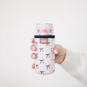 May include: A pink can cooler with a navy blue bow pattern, holding a can of Bubbl'r antioxidant drink. The cooler has a black top and the words "Frederick Family" are printed on the side.