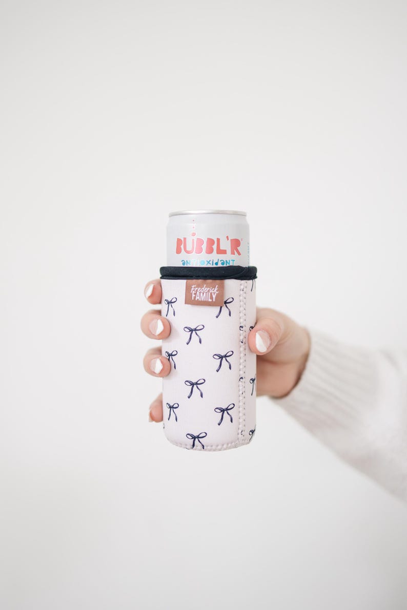 May include: A pink can cooler with a navy blue bow pattern, holding a can of Bubbl'r antioxidant drink. The cooler has a black top and the words "Frederick Family" are printed on the side.