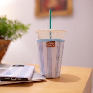 May include: A clear plastic cup with a teal straw inside a light blue and white striped fabric sleeve. The sleeve features a brown rectangular tag with the text Frederick Family Co. printed in white script and a ruler pattern along the side.