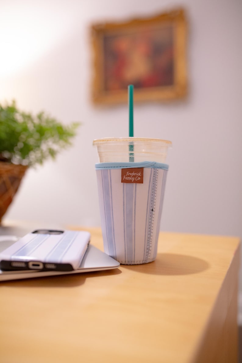 May include: A clear plastic cup with a teal straw inside a light blue and white striped fabric sleeve. The sleeve features a brown rectangular tag with the text Frederick Family Co. printed in white script and a ruler pattern along the side.