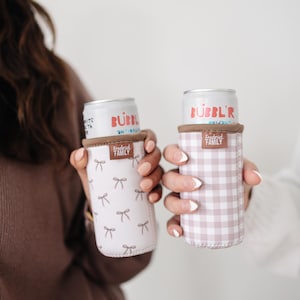 May include: Two beverage can coolers, one with a white background and a pattern of small gray bows, the other with a pink and white gingham pattern. Both have a brown trim and the text "Frederick Family". Cans read "Bubbl'r".