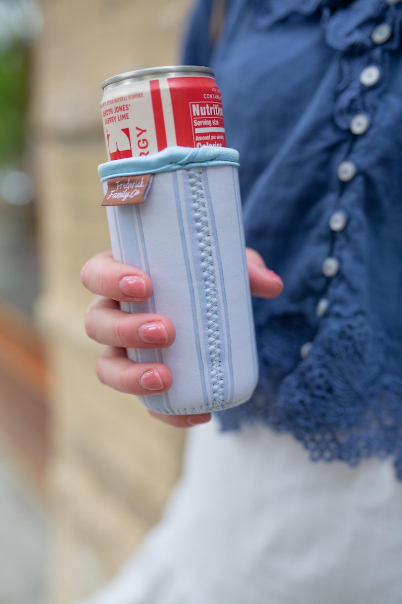 May include: A white and light blue striped beverage insulator with a light blue trim, holding a red and white can. The insulator has a brown tag that reads "Frederick Family Co."