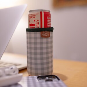 May include: A red and white can of Cherry Lime flavored energy drink sits atop a gray and white checkered can cooler. The cooler has a green rim and a brown leather tag that reads "Fredrick Family Co."