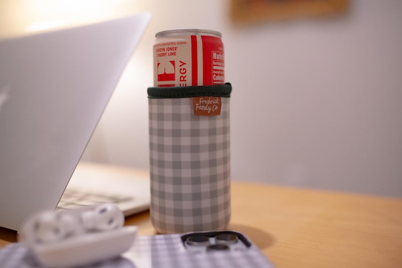 May include: A red and white can of Cherry Lime flavored energy drink sits atop a gray and white checkered can cooler. The cooler has a green rim and a brown leather tag that reads "Fredrick Family Co."