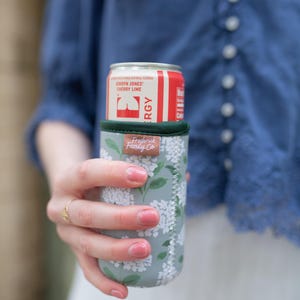 May include: A red and white can of Jordyn Jones Cherry Lime energy drink in a floral print can cooler. The cooler is light green with white flowers and the brand name "Frederick Family Co."