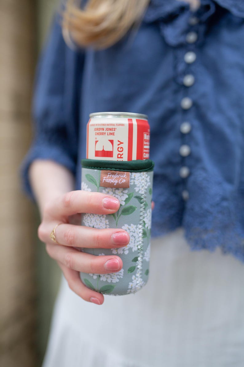 May include: A red and white can of Jordyn Jones Cherry Lime energy drink in a floral print can cooler. The cooler is light green with white flowers and the brand name "Frederick Family Co."