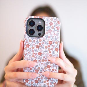 May include: A smartphone case featuring a dense, repeating pattern of small flowers in shades of terracotta, dusty pink, and muted gray on a white background. The case has a black camera cutout frame with three circular lens openings.