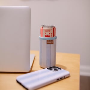 May include: A white and light blue striped can cooler with a red and white can inside. A matching striped phone case is in the foreground. The cooler has a brown leather tag that says "Tropical Family Co."