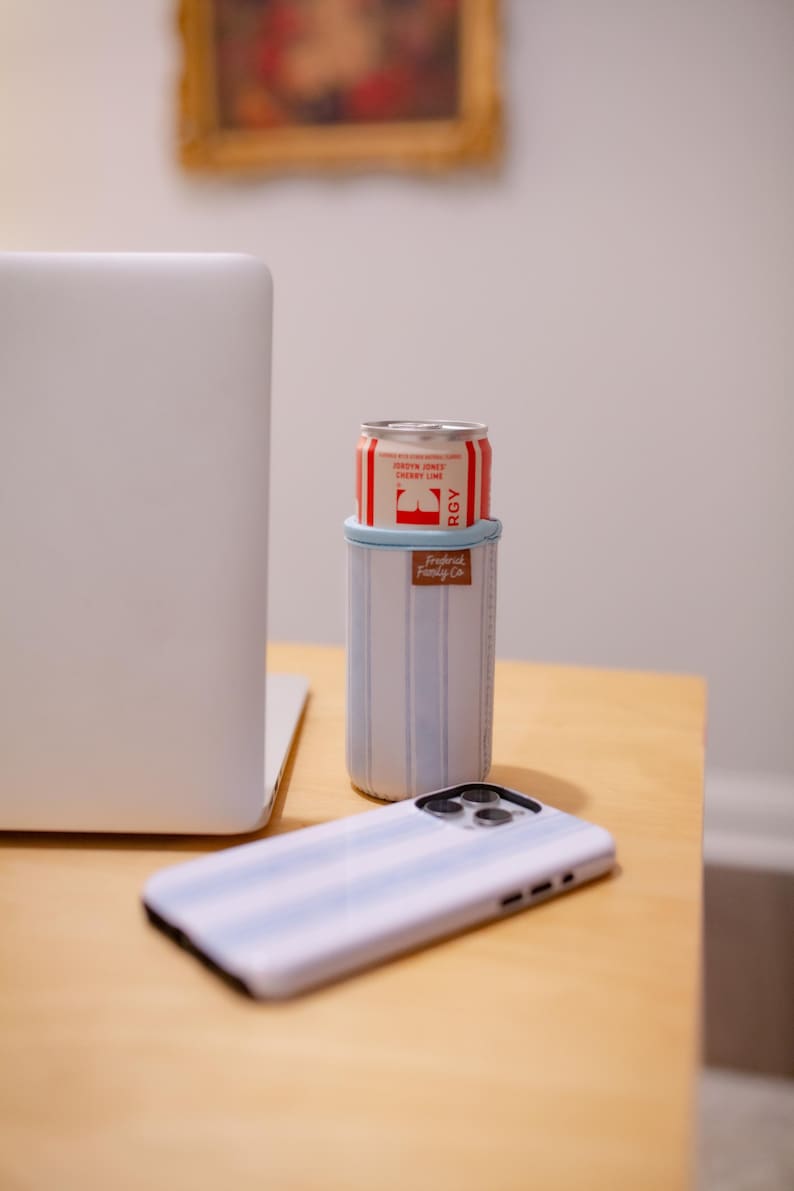 May include: A white and light blue striped can cooler with a red and white can inside. A matching striped phone case is in the foreground. The cooler has a brown leather tag that says "Tropical Family Co."