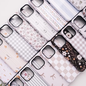 May include: An assortment of phone cases with various patterns, including floral, striped, gingham, and bow designs. The cases feature a black camera cutout and a white or black border, showcasing a range of colors like pink, gray, and brown.