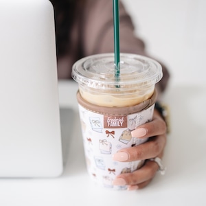 May include: A clear plastic iced coffee cup with a dark green straw, encased in a white fabric sleeve featuring a repeating pattern of small brown bows and books. A small brown tag on the sleeve displays the text Frederick FAMILY in white letters.