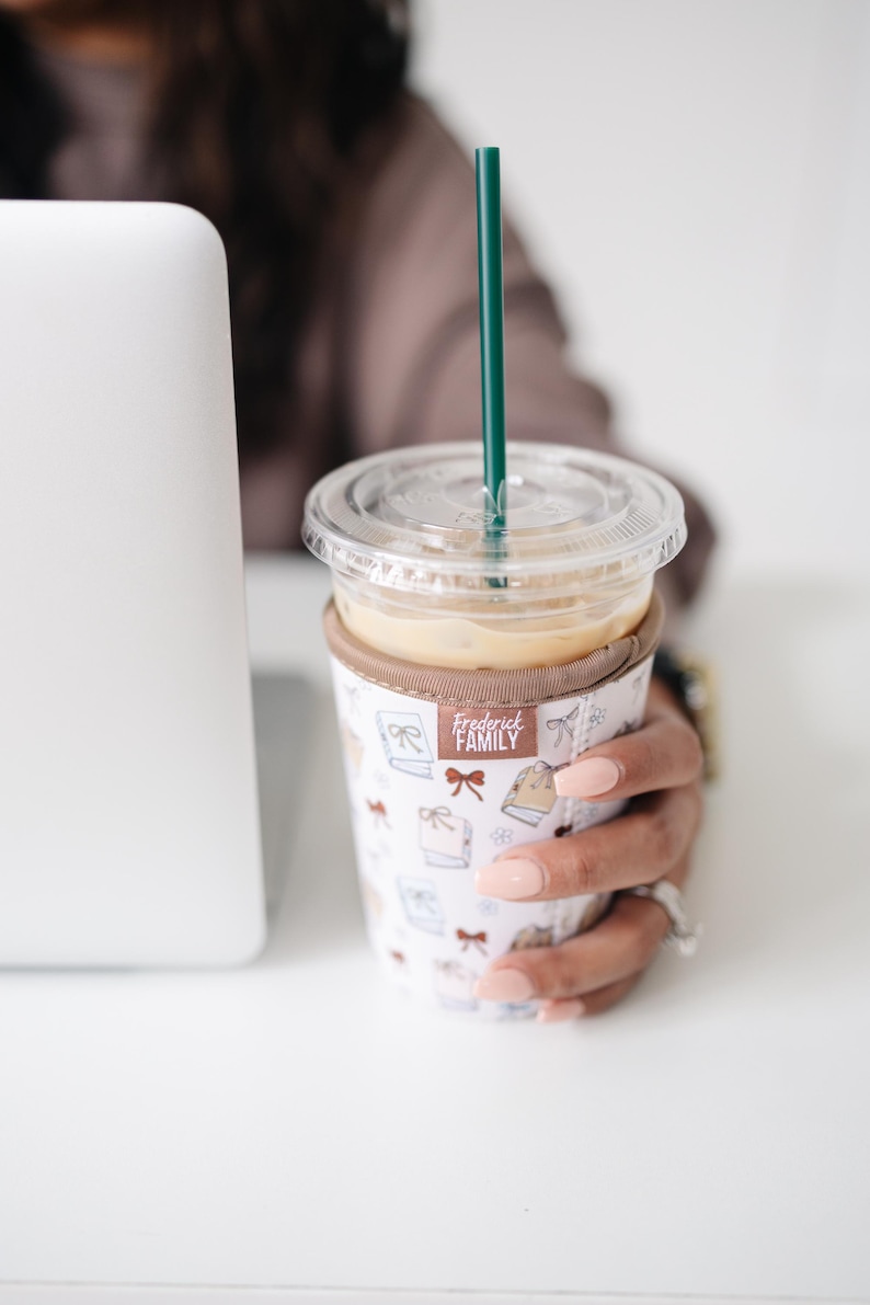 May include: A clear plastic iced coffee cup with a dark green straw, encased in a white fabric sleeve featuring a repeating pattern of small brown bows and books. A small brown tag on the sleeve displays the text Frederick FAMILY in white letters.