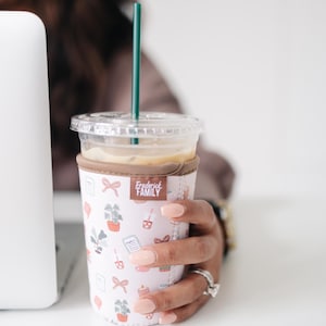 May include: A clear plastic iced coffee cup with a dark green straw, encased in a light pink neoprene sleeve featuring a repeating pattern of small icons including bows, potted plants, candles, and a brown label reading Frederick FAMILY.