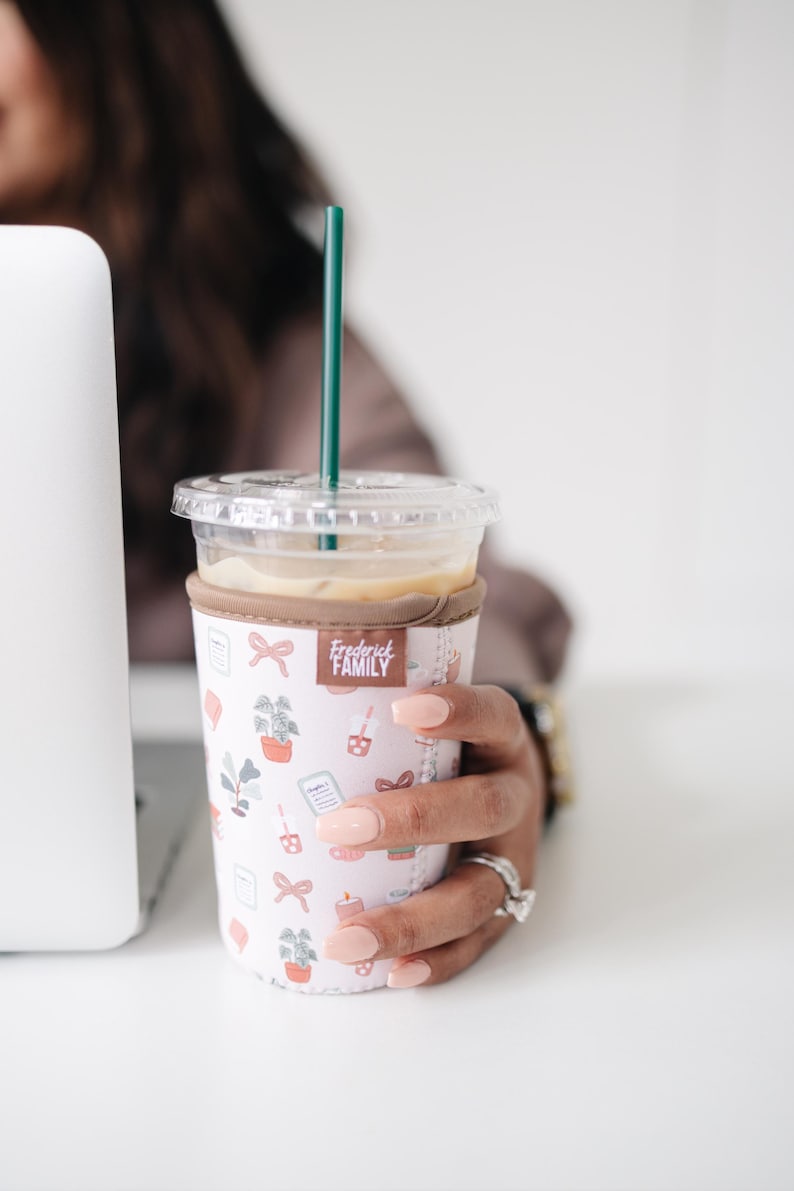 May include: A clear plastic iced coffee cup with a dark green straw, encased in a light pink neoprene sleeve featuring a repeating pattern of small icons including bows, potted plants, candles, and a brown label reading Frederick FAMILY.