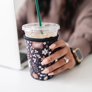 May include: A black neoprene iced coffee cup sleeve featuring a floral pattern in shades of white, peach, and muted pink. The sleeve has a rectangular brown tag with the white text Frederick FAMILY and includes a clear plastic lid and green straw.