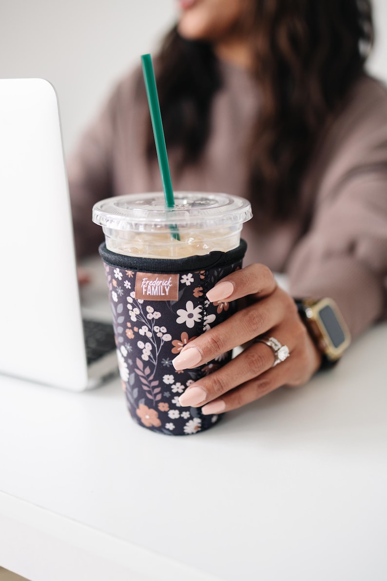 May include: A black neoprene iced coffee cup sleeve featuring a floral pattern in shades of white, peach, and muted pink. The sleeve has a rectangular brown tag with the white text Frederick FAMILY and includes a clear plastic lid and green straw.