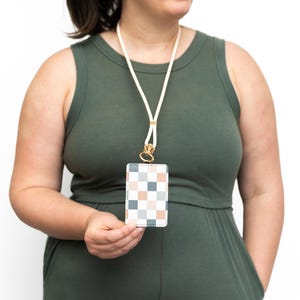 Puede incluir: Un cord&oacute;n blanco con un cierre dorado sostiene un tarjetero rectangular. El tarjetero presenta un patr&oacute;n de cuadros en tonos azules, melocot&oacute;n y blanco. La persona de la imagen lleva un mono verde.