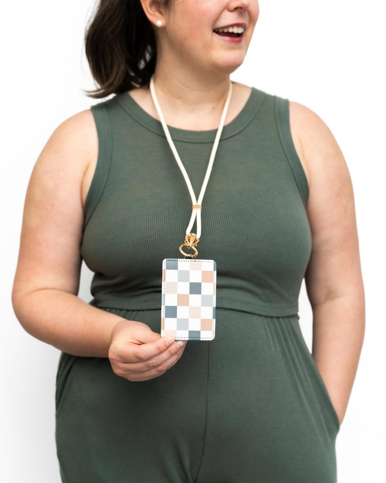Puede incluir: Un cord&oacute;n blanco con un cierre dorado sostiene un tarjetero rectangular. El tarjetero presenta un patr&oacute;n de cuadros en tonos azules, melocot&oacute;n y blanco. La persona de la imagen lleva un mono verde.