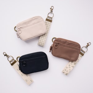 May include: Four small, rectangular coin purses in cream, navy, and brown, each with a zipper closure and a matching wrist strap. The straps feature various patterns, including a gingham and floral design. The purses have a metal ring and clasp.