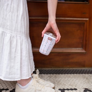 May include: A cylindrical glass tumbler featuring a floral pattern with vertical pink and green stripes, small orange flowers, and green leaves. The tumbler has a clear lid and a small rectangular brown label with white text near the top rim.