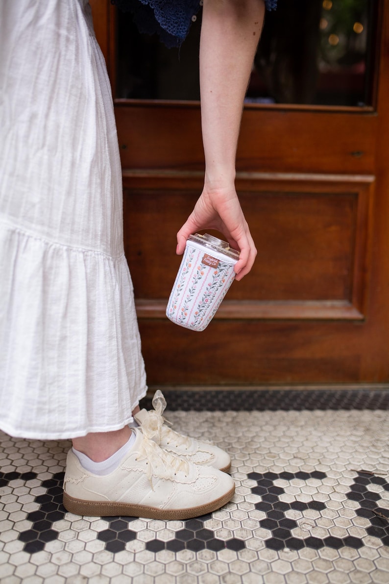 May include: A cylindrical glass tumbler featuring a floral pattern with vertical pink and green stripes, small orange flowers, and green leaves. The tumbler has a clear lid and a small rectangular brown label with white text near the top rim.
