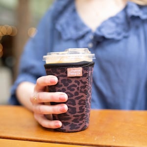 May include: A neoprene cup sleeve featuring a dark brown and black leopard print pattern. The sleeve has a small rectangular tan fabric tag near the top rim with the text "Frosted Farley Co" printed in white. The sleeve fits around a clear plastic cup.