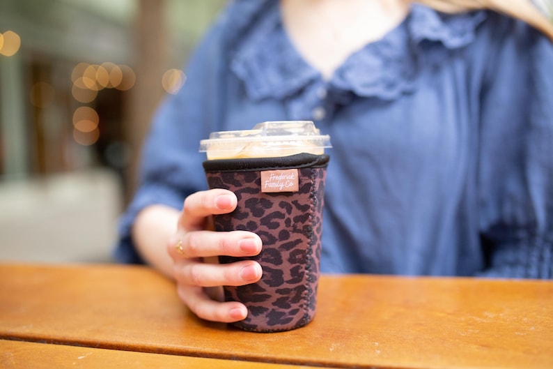 May include: A neoprene cup sleeve featuring a dark brown and black leopard print pattern. The sleeve has a small rectangular tan fabric tag near the top rim with the text "Frosted Farley Co" printed in white. The sleeve fits around a clear plastic cup.