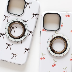 May include: Two white phone cases with different designs. One case has a pattern of black bows and the other has a pattern of red cherries. Both cases have a gold ring holder.