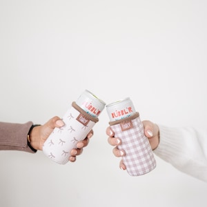 May include: Two beverage insulators, one with a light pink bow pattern and the other with a pink gingham pattern, each holding a can. The cans have the word "BUBBL'R" printed on them. The insulators have a brown band at the top and the words "FRIENDLY FAMILY" printed on them.