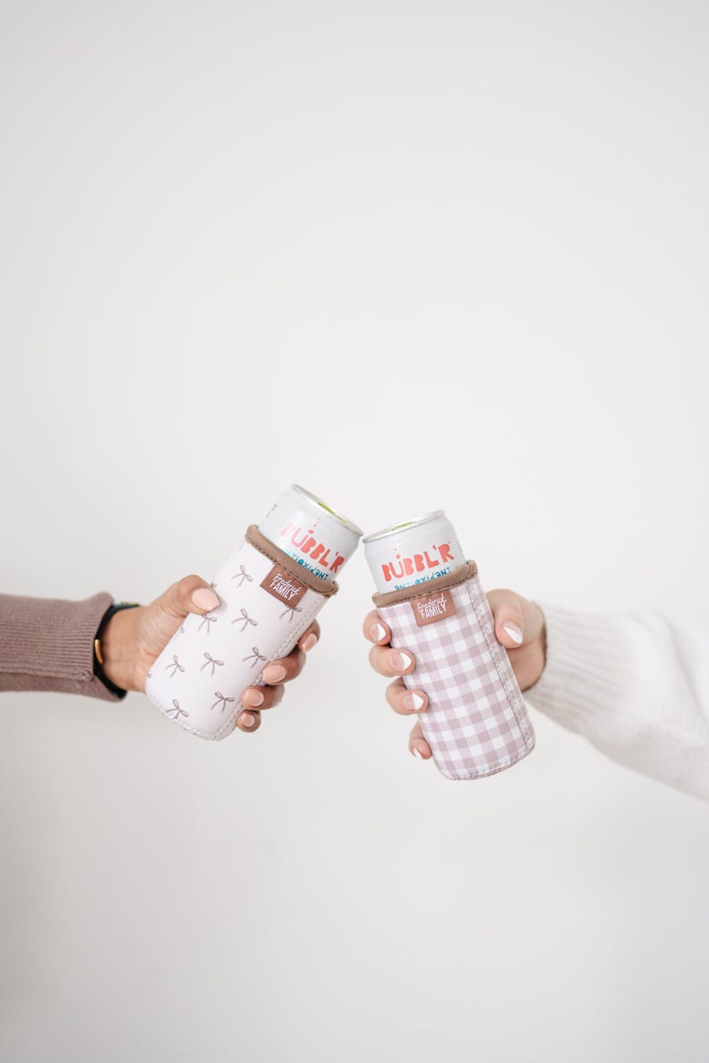 May include: Two beverage insulators, one with a light pink bow pattern and the other with a pink gingham pattern, each holding a can. The cans have the word "BUBBL'R" printed on them. The insulators have a brown band at the top and the words "FRIENDLY FAMILY" printed on them.