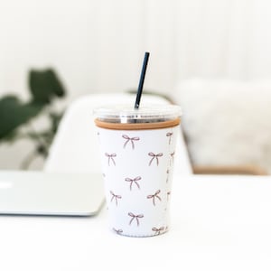 May include: A white insulated cup sleeve featuring a repeating pattern of small brown bows. The sleeve has a tan zipper closure along the side and is fitted around a clear plastic tumbler with a clear lid and a thin black straw inserted in the top.