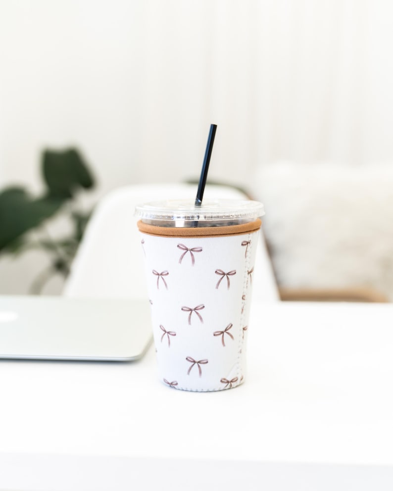 May include: A white insulated cup sleeve featuring a repeating pattern of small brown bows. The sleeve has a tan zipper closure along the side and is fitted around a clear plastic tumbler with a clear lid and a thin black straw inserted in the top.