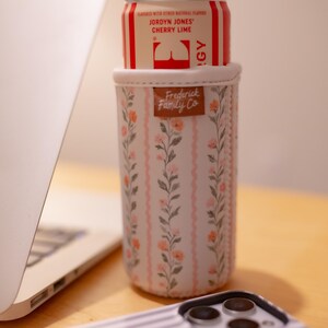May include: A floral insulated can cooler with a can of Jordyn Jones Cherry Lime soda. The cooler has a pink and white floral pattern with the words "Frederick Family Co" on a brown leather tag. A laptop and phone are in the background.