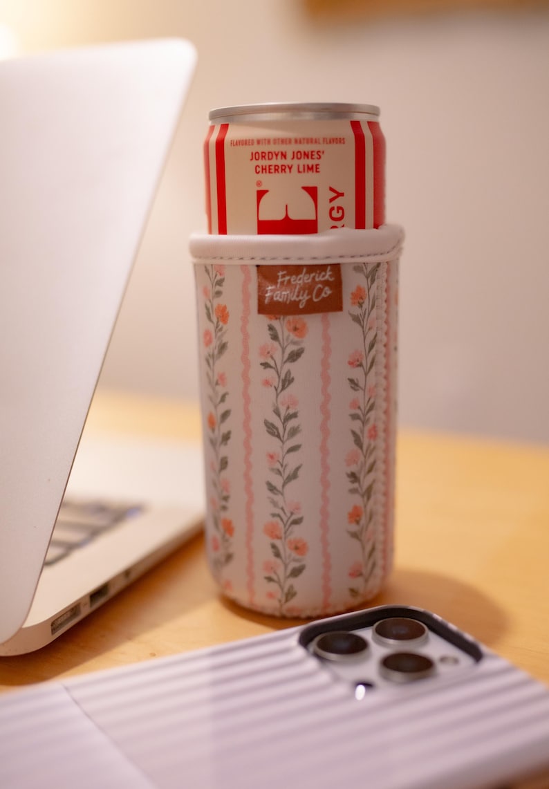May include: A floral insulated can cooler with a can of Jordyn Jones Cherry Lime soda. The cooler has a pink and white floral pattern with the words "Frederick Family Co" on a brown leather tag. A laptop and phone are in the background.