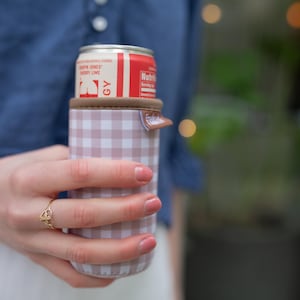 May include: A can of cherry lime soda in a pink and white checkered can cooler. The can cooler has a brown top and a small tab. The soda can is red and white with the words "Cherry Lime" visible.