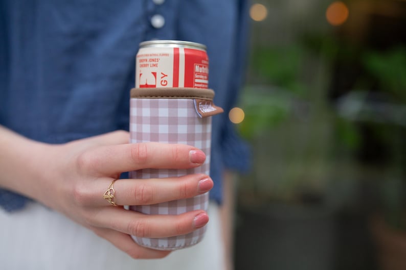 May include: A can of cherry lime soda in a pink and white checkered can cooler. The can cooler has a brown top and a small tab. The soda can is red and white with the words "Cherry Lime" visible.