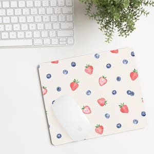Puede incluir: Una alfombrilla de ratón blanca con un patrón repetitivo de fresas rojas y arándanos azules sobre un fondo blanco.