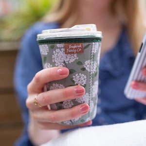 May include: A sage green neoprene coffee cup sleeve featuring a white floral pattern and a small brown rectangular tag with the text Frederick Family Co. The sleeve has a dark green top trim and visible white stitching along the side seam.