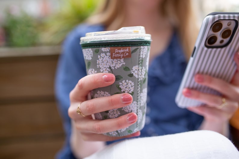 May include: A sage green neoprene coffee cup sleeve featuring a white floral pattern and a small brown rectangular tag with the text Frederick Family Co. The sleeve has a dark green top trim and visible white stitching along the side seam.
