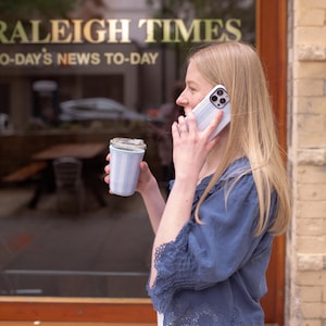 May include: A light blue and white striped reusable coffee cup with a clear lid, held alongside a white smartphone with a matching light blue and white striped case, featuring a blue blouse with scalloped lace trim and a white skirt.